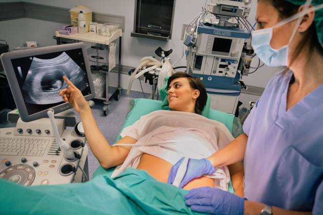 Female doctor in sterile mask and blue glove using ultrasound scanner while examining cheerful pregnant woman pointing at the screen in hospital, Model released, Property released Copyright: xSixAndxSix MR