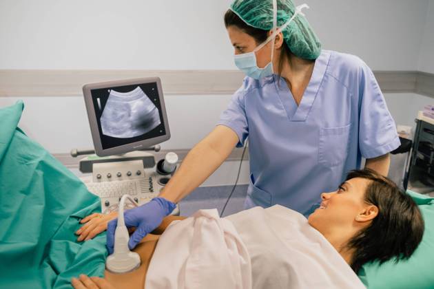Female doctor in sterile mask and blue glove using ultrasound scanner while examining cheerful pregnant woman in hospital, Model released, Property released Copyright: xSixAndxSix MR