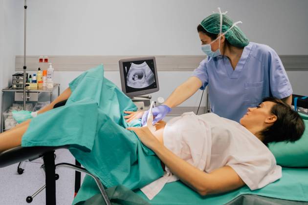 Female doctor in sterile mask and blue glove using ultrasound scanner while examining cheerful pregnant woman in hospital, Model released, Property released Copyright: xSixAndxSix MR