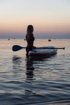 Side view of of unrecognizable female surfer standing near paddleboard and rowing against spectacular sun in sunset sky Copyright: xVictorxPrietox Model Released