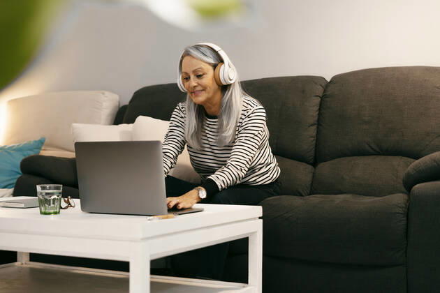 Smiling woman wearing headphones using laptop while sitting on sofa at home model released Symbolfoto property released ERRF04773