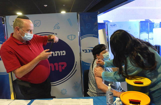 A man takes a photo of his wife as she receive the second shot of the Pfizer- BioNTech COVID-19 vaccine in an Israeli vaccination center in Jerusalem, on Tuesday, January 12, 2021. Israel will expand the vaccination drive inoculating teachers and people over 50 years on Wednesday.  JER2021011210 DEBBIExHILL