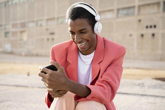 IMAGO Creative: Clickbait, Modetrends Happy man wearing headphones using mobile phone while sitting outdoors model released Symbolfoto AFVF07894