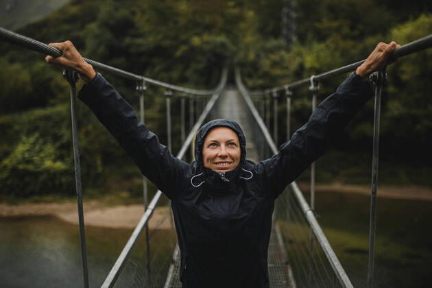 IMAGO Creative: Ende des Sommers Creative Highlights Symbolbilder Smiling woman wearing raincoat with arms raised standing on suspension bridge over Sella river in forest model released Symbolfoto DMGF00421