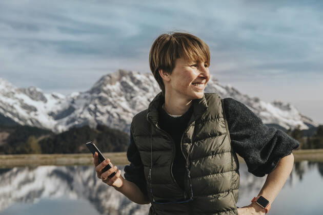 Smiling woman with smart phone looking away while standing at Prinzensee, Hochkoenig massif, Salzburger Land, Austria model released Symbolfoto MFF07221