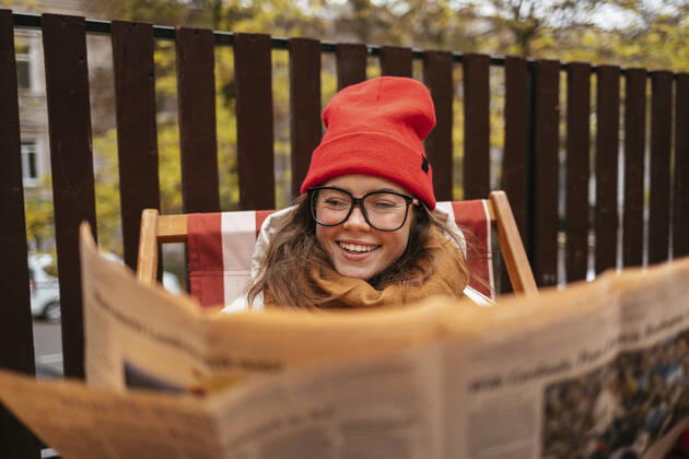 IMAGO Creative: Kulturelle Folgen der Pandemie: Gesundheitsbewußtsein und Alleinsein Smiling woman wearing knit hat and eyeglasses reading newspaper while sitting at sidewalk cafe model released Symbolfoto OYF00340