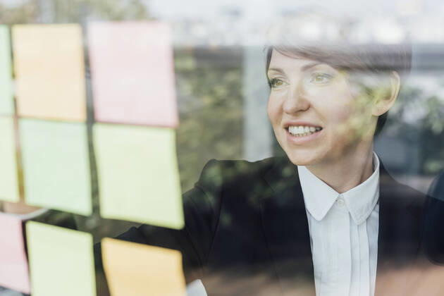Smiling female entrepreneur looking at adhesive notes on glass wall model released Symbolfoto property released MEUF02264