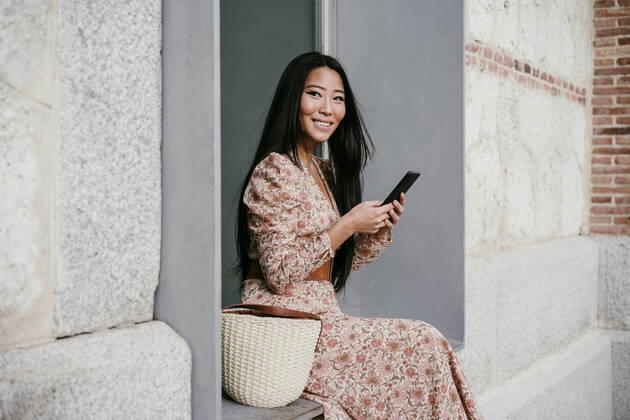 Smiling woman with smart phone looking away while sitting on window sil model released Symbolfoto EBBF03196
