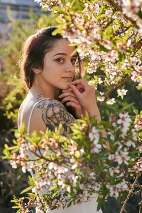 Young female with tattooed arm wearing white dress and standing in flowers of tree looking at camera, Model released, Property released Copyright: xCristianxBlazquezx MR