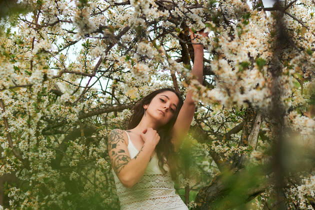 Young female with tattooed arm wearing white dress and standing in flowers of tree looking at camera, Model released, Property released Copyright: xCristianxBlazquezx MR