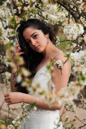 Young female with tattooed arm wearing white dress and standing in flowers of tree looking at camera, Model released, Property released Copyright: xCristianxBlazquezx MR