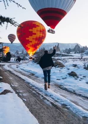 Back view of unrecognizable man in warm clothes with arm raised running to vivid colored air balloon flying up into gray sky over foggy snowy mountain ridge in Turkey, Model released Copyright: xCarlosxForteax MR