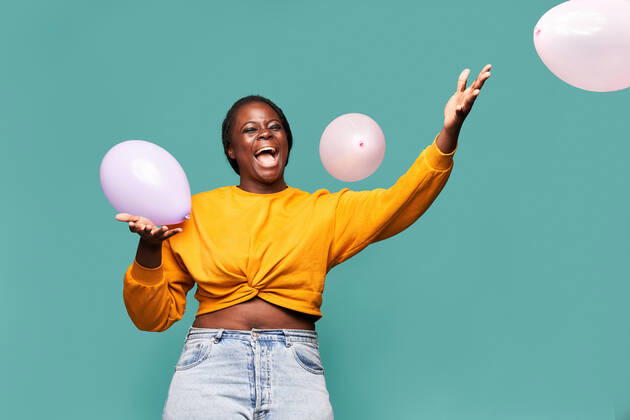Excited African American female in jeans and yellow top standing near falling balloons against blue background in studio Copyright: xManuelxRuizx Model Released