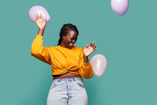 Excited African American female in jeans and yellow top standing near falling balloons against blue background in studio Copyright: xManuelxRuizx Model Released