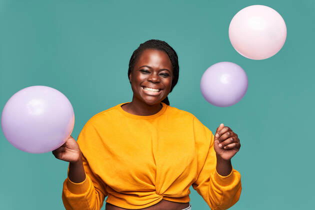 Excited African American female in jeans and yellow top standing near falling balloons against blue background in studio Copyright: xManuelxRuizx Model Released