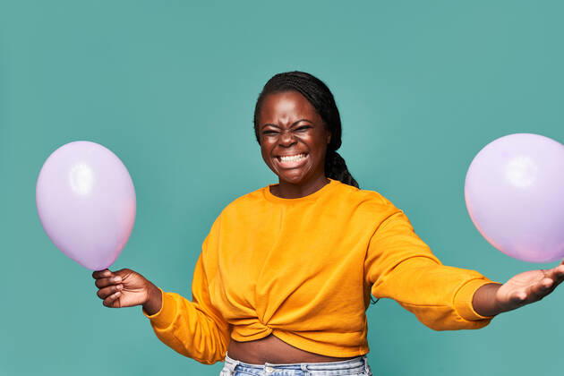 Excited African American female in jeans and yellow top standing near falling balloons against blue background in studio Copyright: xManuelxRuizx Model Released