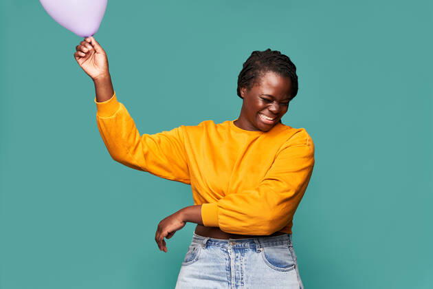 Excited African American female in jeans and yellow top standing near falling balloons against blue background in studio Copyright: xManuelxRuizx Model Released