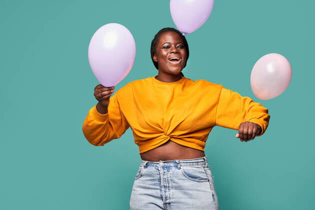 Excited African American female in jeans and yellow top standing near falling balloons against blue background in studio Copyright: xManuelxRuizx Model Released