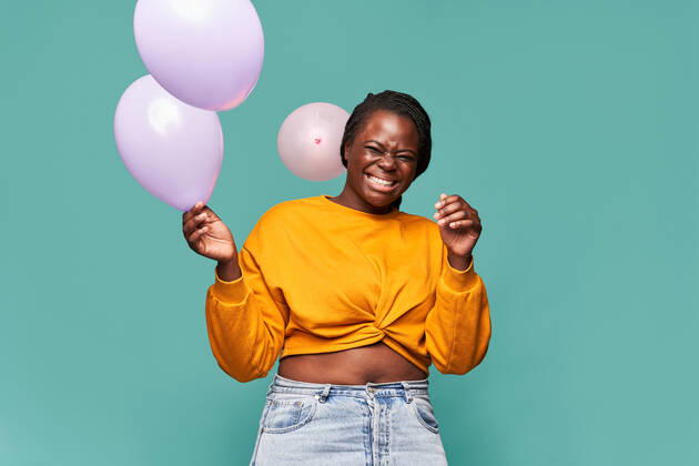 Excited African American female in jeans and yellow top standing near falling balloons against blue background in studio Copyright: xManuelxRuizx Model Released