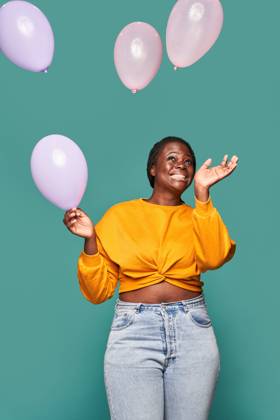 Excited African American female in jeans and yellow top standing near falling balloons against blue background in studio Copyright: xManuelxRuizx Model Released