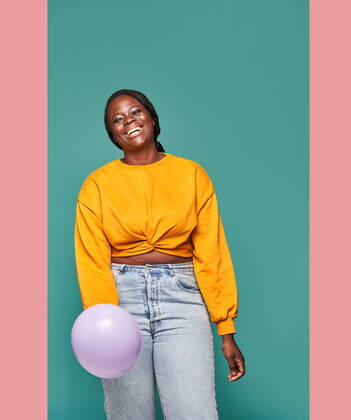 Excited African American female in jeans and yellow top standing near falling balloons against blue background in studio Copyright: xManuelxRuizx Model Released