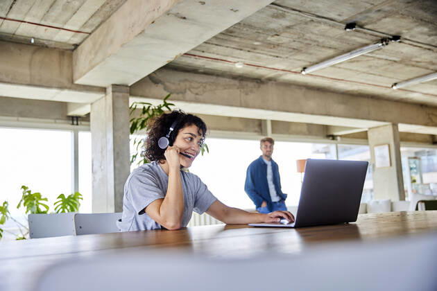 Smiling woman wearing headphones using laptop with boyfriend in background at home apartment model released Symbolfoto property released FMKF07169
