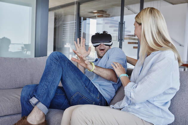 Excited man wearing virtual reality simulator sitting by cheerful woman in living room model released Symbolfoto property released RORF02804