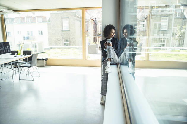 Businesswoman holding coffee cup while sitting on window sill at office model released Symbolfoto property released UUF23526