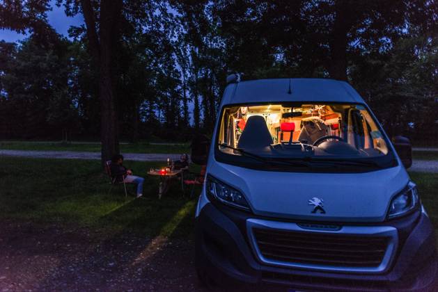 Camper sitzen zur blauen Stunde an einem lauen Sommerabend vor ihrem Camper auf Campingstühlen. Baden Württemberg, Deutschland. Mit einem Campervan unterwegs