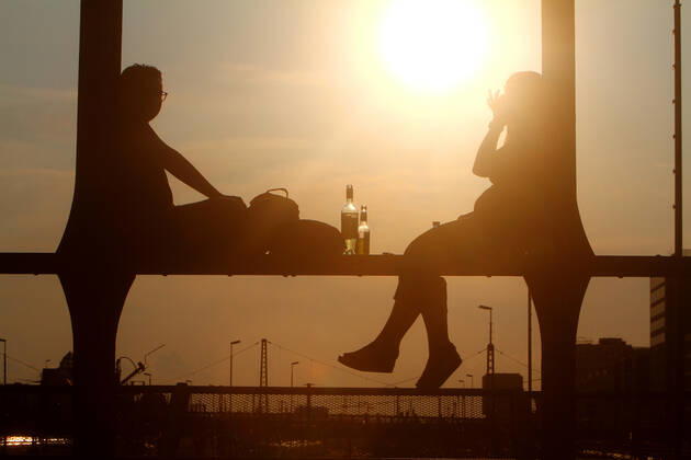 Ein Paar sitzt im Gegenlicht der untergehenden Sonne auf einem Träger der Hackerbrücke in München mit einem Feierabendbier und unterhält sich, Bayern, Deutschland.