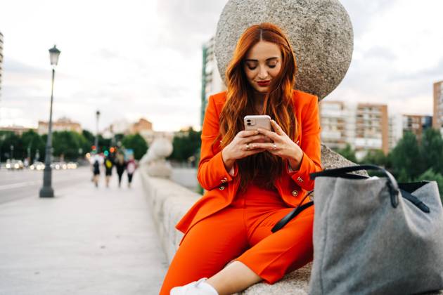 Happy stylish female with red hair and in vibrant orange suit sitting on stone border in city and messaging on cellphone JakeJakab_MariaPortraits_33.jpg Copyright: xJakexJakabx Model Released