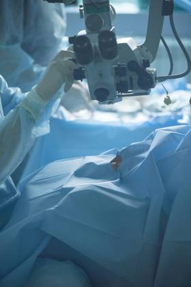 Adult female doctor in sterile mask and ornamental medical cap looking through surgical microscope against crop coworker in hospital, Model released SergeyMironov_Surgery_2.jpg Copyright: xSergeyxMironovx MR