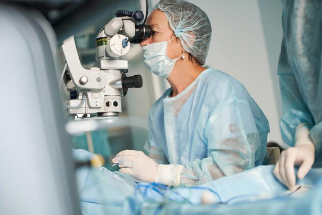 Adult female doctor in sterile mask and ornamental medical cap looking through surgical microscope against crop coworker in hospital, Model released SergeyMironov_Surgery_32.jpg Copyright: xSergeyxMironovx MR