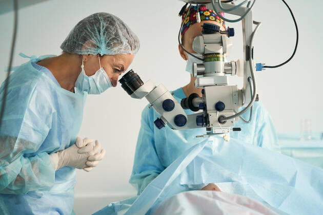 Adult female doctor in sterile mask and ornamental medical cap looking through surgical microscope against crop coworker in hospital, Model released SergeyMironov_Surgery_36.jpg Copyright: xSergeyxMironovx MR