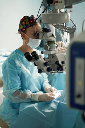 Adult female doctor in sterile mask and ornamental medical cap looking through surgical microscope against crop coworker in hospital, Model released SergeyMironov_Surgery_47.jpg Copyright: xSergeyxMironovx MR