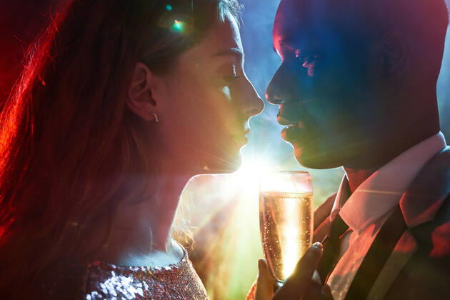 Side view of crop couple with glass on champagne in moment of kiss against shiny light ray looking at each other during party ManuelRuiz_NewYearsEvePartyCouple_32.jpg Copyright: xManuelxRuizx Model Released