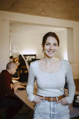 Portrait of smiling businesswoman with hands in pockets while colleague working at desk in office Germany, Berlin, model released, property released Copyright: xMaskotx , MA90121