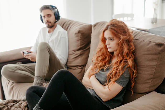 Full body of sad redhead female sitting on comfortable couch near boyfriend playing interesting videogame in light living room at home Copyright: xSunsixAlbetsx Model Released
