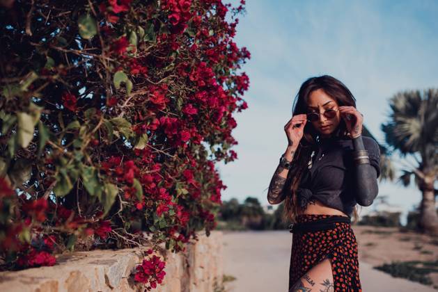 Confident stylish tattooed female with nose piercing and bindi touching sunglasses with closed eyes on the street against blurred shrub with vinous flowers, Model released RemediosPuerta_BohoBea_3 Copyright: xRemediosxPuertax MR
