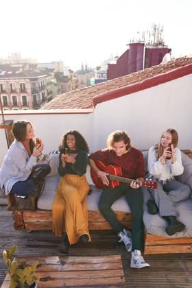 IMAGO Kreativ: Urbane Kultur - Sommer auf den D?chern Full length happy young multiracial friends in casual outfits playing guitar and enjoying beer while gathering together on comfy sofa on terrace, Model released, Property released ManuelRuiz_FlatmatesMusiciansTerrace_32 Copyright: xManuelxRuizx MR