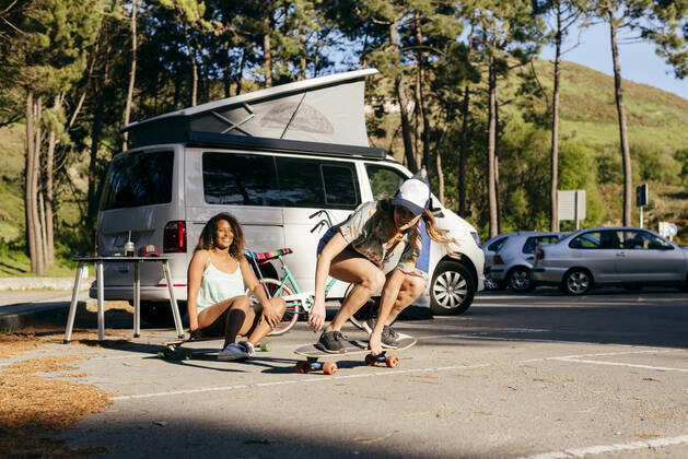 IMAGO Creative, Jahreszeit Sommer, Sommerferien Beautiful skater practicing riding skate board on street with background of woman sitting on skate near minivan in camping, Model released ManuelRuizAlba_CamperGirls_84 Copyright: xManuelxRuizxAlbax MR