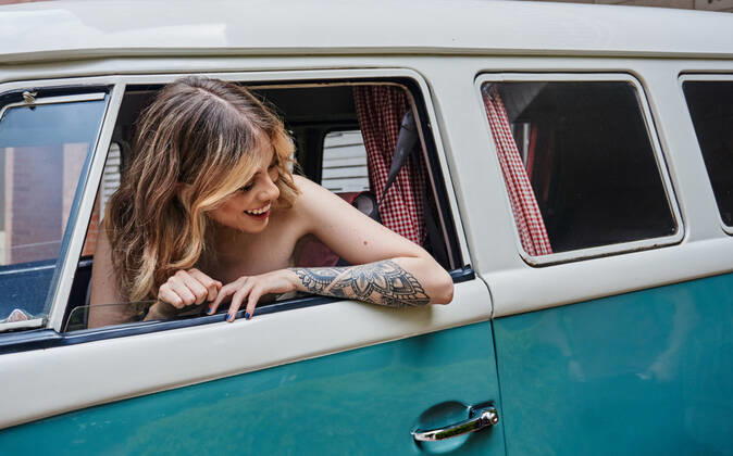 IMAGO Creative, Jahreszeit Sommer, Sommerferien Delighted young female driver with long wavy hair and tattoo on arm smiling and looking away while sitting in vintage van during holidays JoseDeJesusSaldana_ID10013_302921_038 Copyright: xJosexDexJesusxSaldanax Model Released