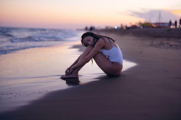 Side view of young woman in white swimwear embracing knees and closing eyes while sitting on beach near sea at sunset, Model released XavierLaiz_ID10743_325282_033 Copyright: xXavierxLaizx MR