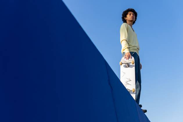 Low angle full body of thoughtful young Asian male skater in casual outfit standing on ramp with skateboard and looking away against blue cloudless sky, Model released DavidMunoz_ID12930_392769_042 Copyright: xDavidxMunozx MR