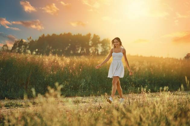 IMAGO Creative: Sommer, Abendspaziergang Glückliche junge Frau, die bei Sonnenuntergang auf einer Wiese entlang der Landstraße spaziert