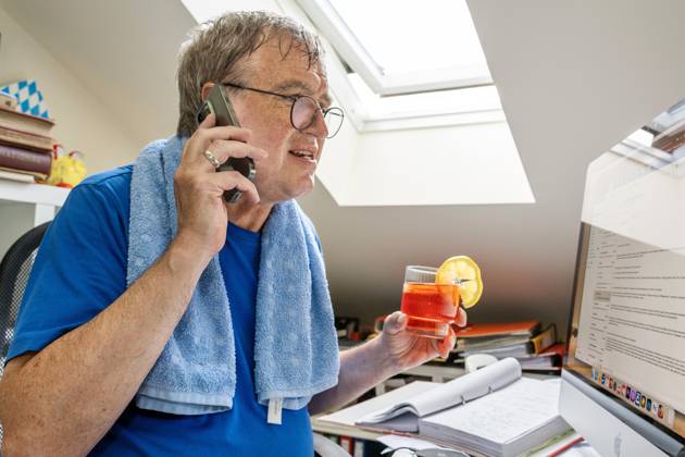 IMAGO Creative, Jahreszeit Sommer, Hitze Mann schwitzt im Home Office, leidet unter Rekordhitze in der Dachwohnung, München, Juli 2023 Deutschland, München, Juli 2023, Mann schwitzt im Home Office, leidet unter Hitze in der Dachwohnung, gönnt sich ein kühles Getränk, Glas Aperol, sitzt unter der Dachschräge, Sonne knallt durch das Fenster ins Büro, Schweiß läuft ihm übers Gesicht, er telefoniert und arbeitet am Computer, Alkohol in der Arbeitszeit, Handtuch liegt um die Schultern, schwitzen, Dachfenster geöffnet, Brille, Wohnung, Bayern,