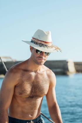 Young fit shirtless man in swimming shorts and hat standing on sailboat enjoying summer vacation against sea and cloudless blue sky Model Released AlvaroMontero_ID16707_510355_005 Copyright: xAlvaroxMonterox RECORD DATE NOT STATED