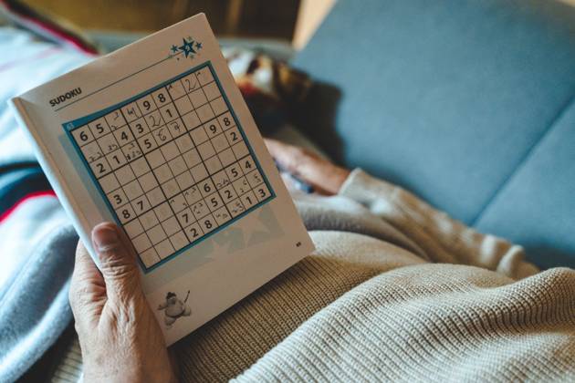 An old woman lies on a sofa in the living room and plays Sudoku in a book with a pen