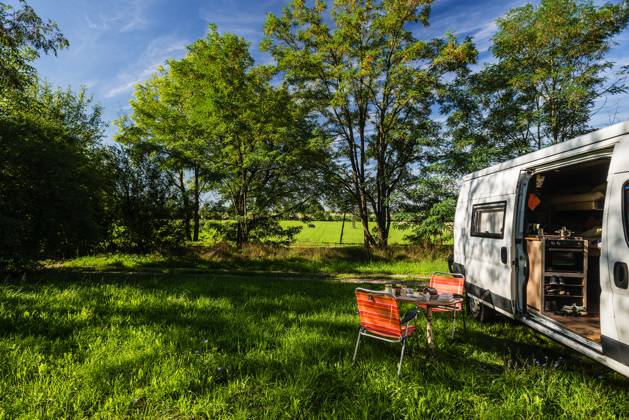 Ein Camper steht zwischen Bäumen an einem sonnigen Sommertag auf einer Wiese im Grünen, ein gedeckter Frühstückstisch mit Stühlen steht vor der Schiebetür des Campers. Regensburg, Bayern, Deutschland. In der Altstadt von Regensburg