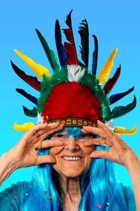IMAGO Creative, Gesellschaft, Fasching Portrait of smiling senior lady blue in wig with confetti wearing Indian headdress with colorful feathers while standing and touching face against blue background and looking at camera Model Released SergioVictorVega_ID17016_518845_023 Copyright: xSergioxVictorxVegax
